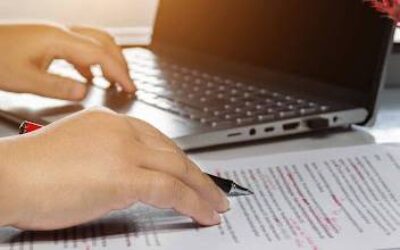 An image of a laptop and a person proofreading a stack of papers which represents knowing the disparities between editing and proofreading is crucial in grant writing. These stages may overlap, but they serve distinct purposes. Both are vital for refining, organizing, and submitting a polished final document. Understanding their nuances allows grant writers to improve proposal quality and boost success rates.